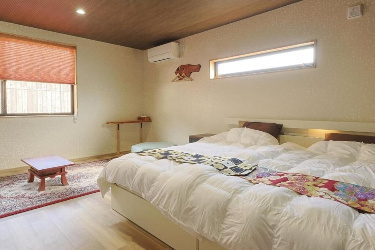A bedroom with a bed, a small wooden stool, a window with blinds, and a wall-mounted light fixture.