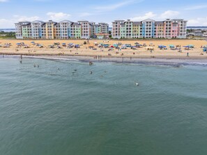 Ein Hotel direkt am Strand mit mehreren bunten Gebäuden, einem Sandstrand mit Sonnenschirmen und einem ruhigen Meer.
