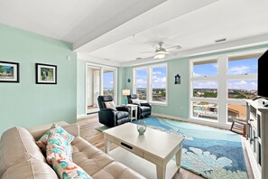 A spacious living room with a large window, a ceiling fan, and a view of the surrounding area. - Book Wapacajune Prop 2 | Sandbridge Vacation Rental (virginia beach)