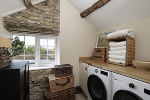 A laundry room with a washing machine, stacked baskets, and a window with a view of greenery. - Top House -  a country house that sleeps 8 guests  in 4 bedrooms (Shipton-under-Wychwood)