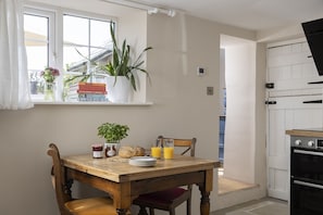 Eine Küche mit einem Holztisch, Stühlen, einem Blumentopf, einem Fenster mit Vorhängen und einer weißen Tür.