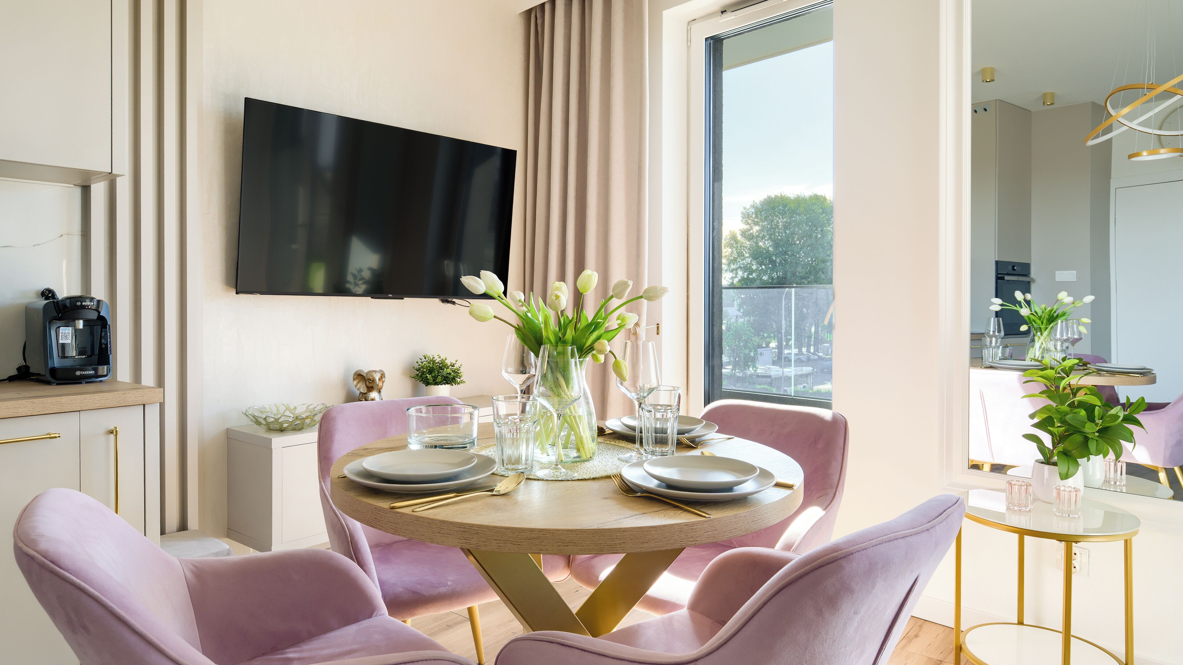 A dining area with a round table, pink chairs, a vase with flowers, and a flat-screen TV mounted on the wall.