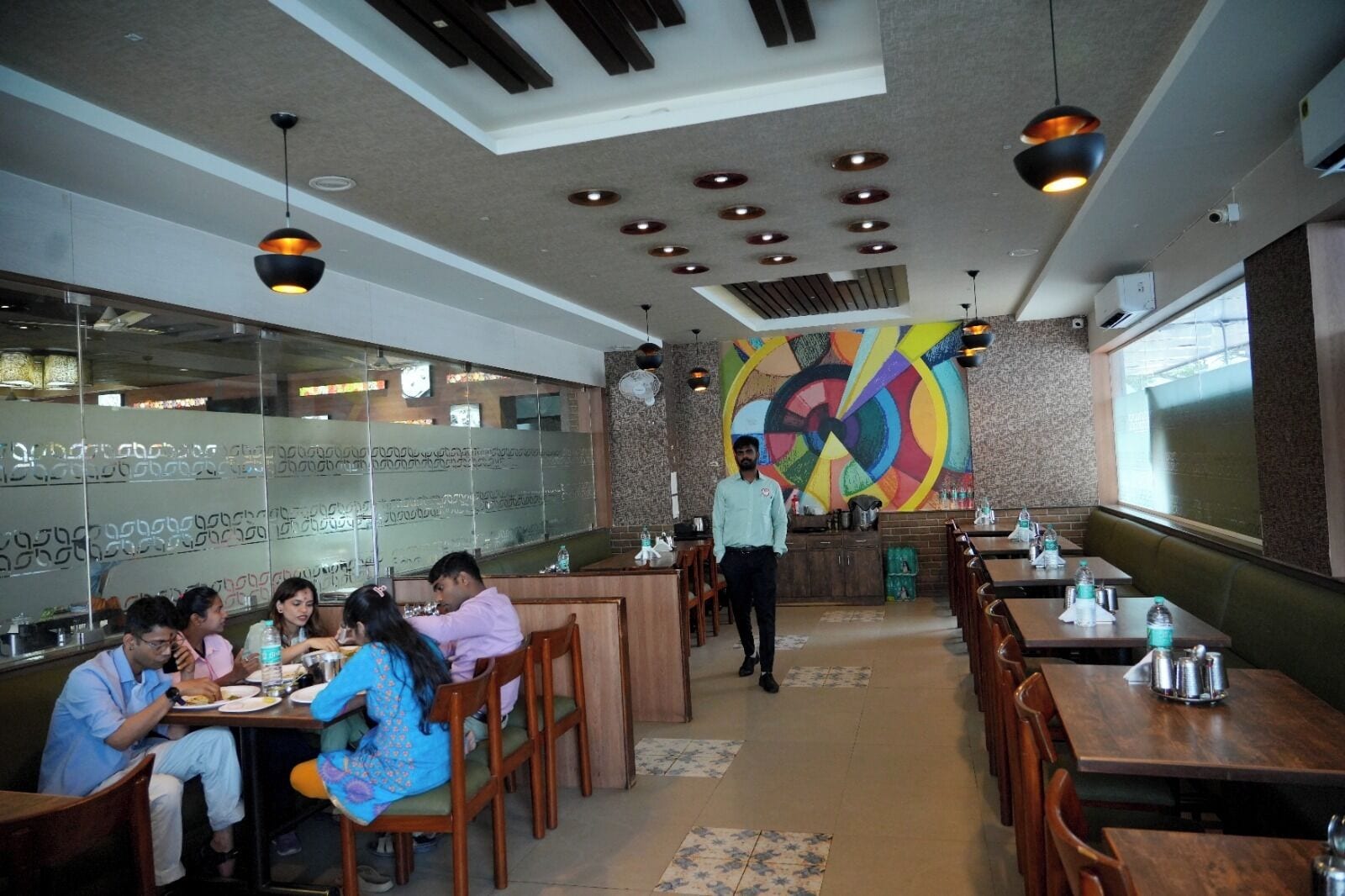 A restaurant interior with wooden tables, a colorful abstract wall art, and a person walking.