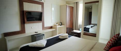 A hotel room with a bed, a TV mounted on the wall, a desk with a chair, a mirror, and a window with curtains.