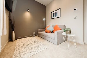 Ein Wohnzimmer mit grauem Sofa, einem gemusterten Teppich, einem kleinen Tisch mit Pflanze und einem Wandbild.