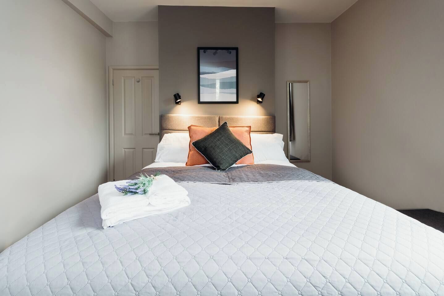 A hotel room with a bed, a headboard, a framed picture above the bed, and a nightstand with a lamp.