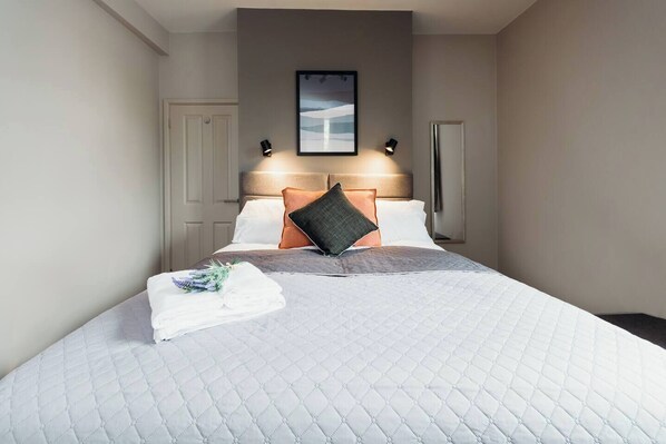 A hotel room with a bed, a headboard, a framed picture above the bed, and a nightstand with a lamp.