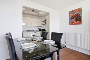 A dining area with a glass table set for four, black chairs, and a kitchen in the background. - #191 Mason Way Executive Stay - Sleeps 6 (West Midlands)
