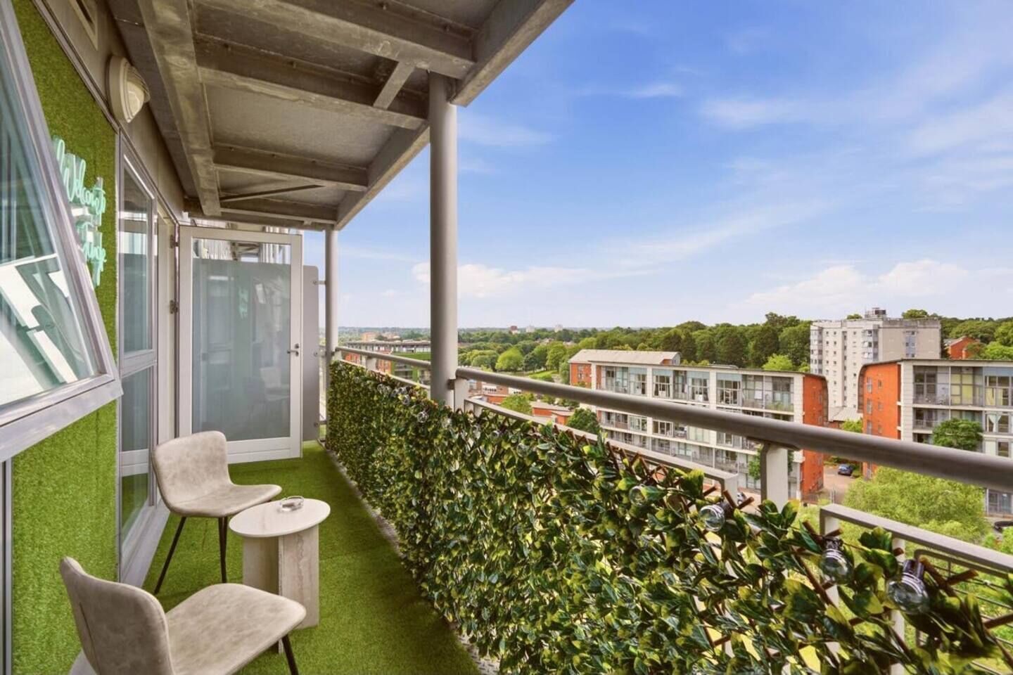 Een balkon met kunstgras, twee stoelen, een tafeltje en uitzicht op gebouwen en groen.