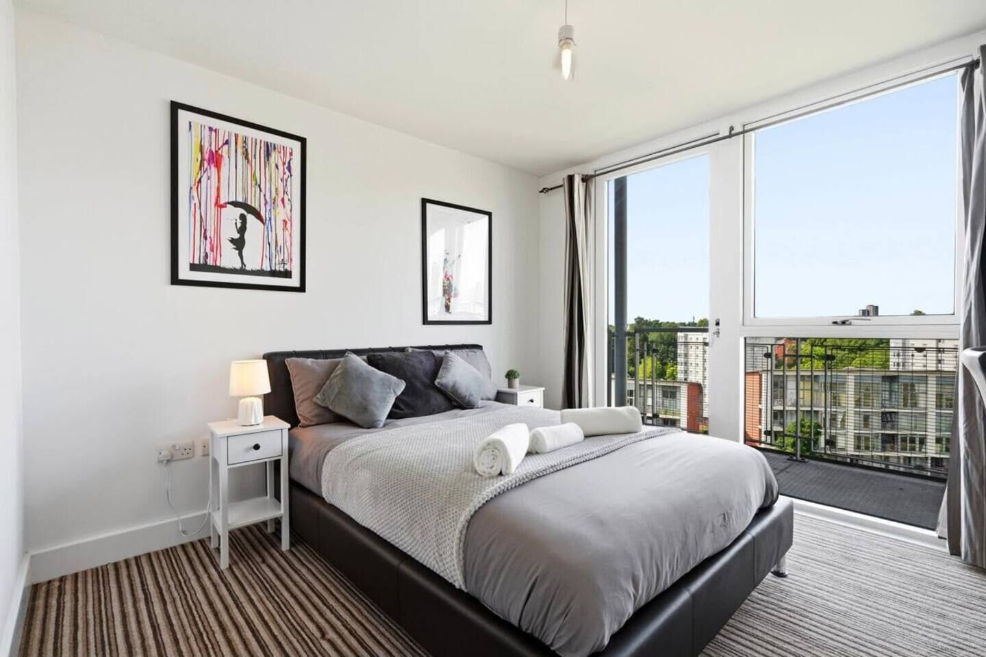 Een moderne slaapkamer met een groot bed, nachtkastje en een raam met uitzicht op gebouwen.