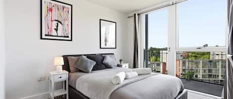 A modern bedroom with a large bed, a nightstand, a bedside table, and a window with a view of buildings.
