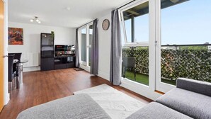 Un soggiorno moderno con divano, angolo da pranzo con tavolo e sedie, zona cucina con TV e ampie porte scorrevoli in vetro che danno su un balcone con vista sulla città.