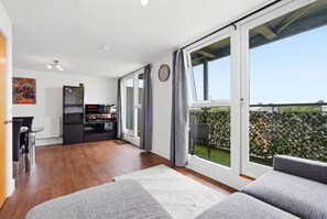 A modern living room with a sofa, a dining area with a table and chairs, a kitchen area with a TV, and large sliding glass doors leading to a balcony with a view of the city. - #191 Mason Way Executive Stay - Sleeps 6 (West Midlands)