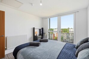 A modern hotel room with a large bed, a desk with a lamp, and a view of the city through a large window. - #191 Mason Way Executive Stay - Sleeps 6 (West Midlands)