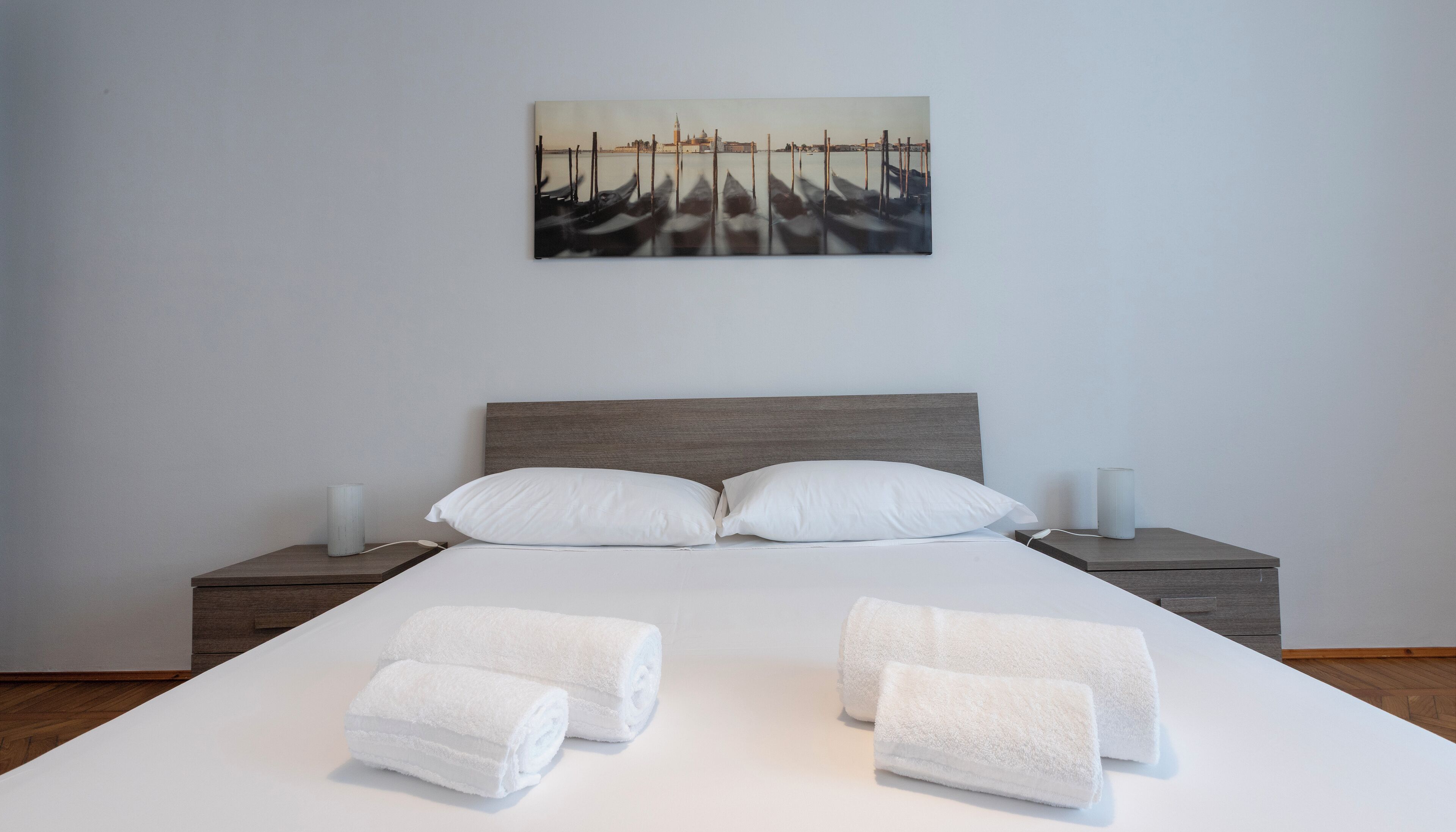 Hotellihuone, jossa on suuri sänky, kaksi yöpöytää, taulu seinällä ja sängyllä rullalle taitetut pyyhkeet.
