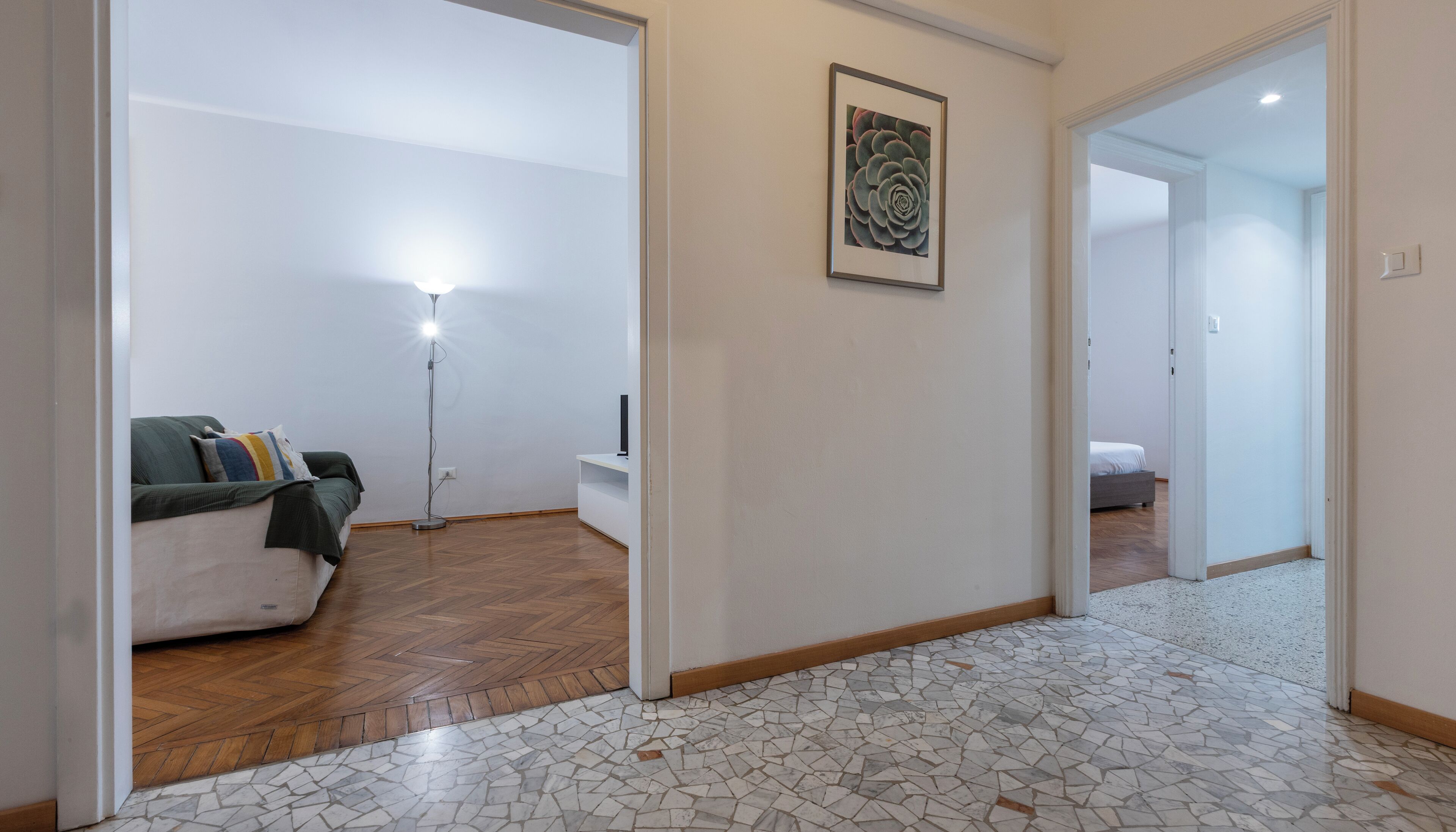 A hallway with a tiled floor, a sofa with cushions, a standing lamp, and a framed picture on the wall.