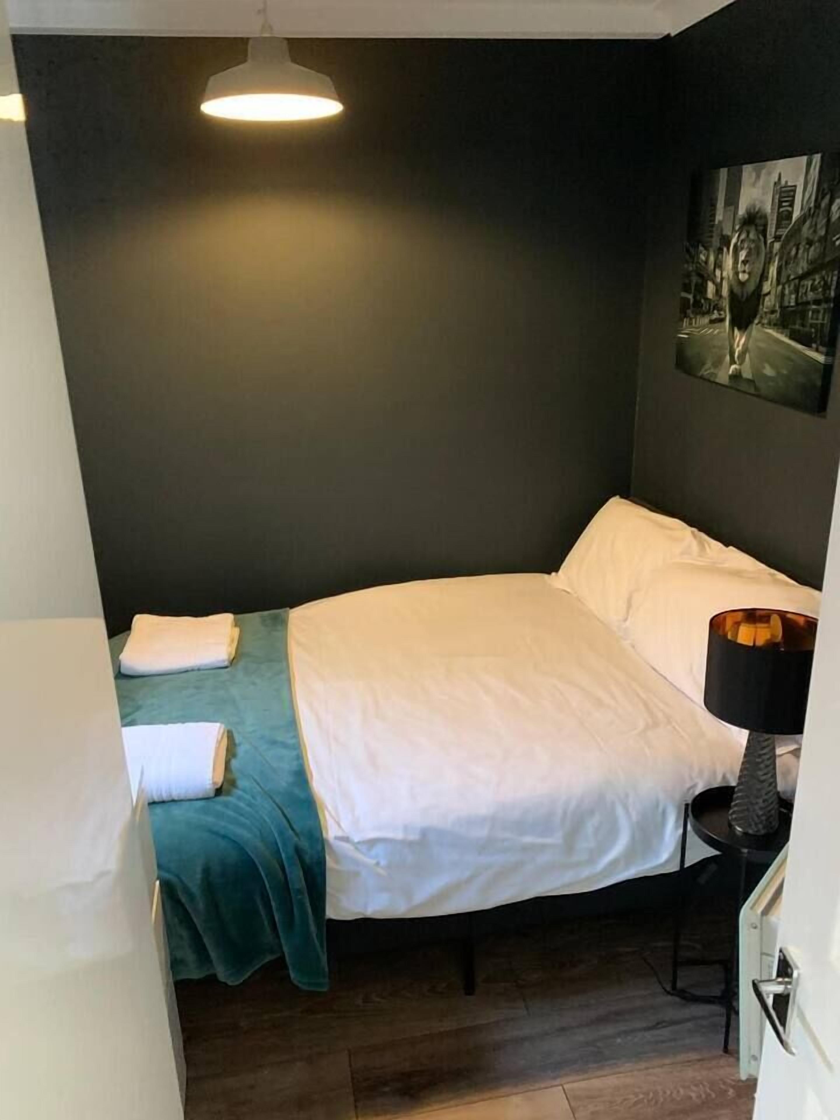 A small, single-bed room with a dark wall, a painting, a bedside lamp, and a wooden floor.
