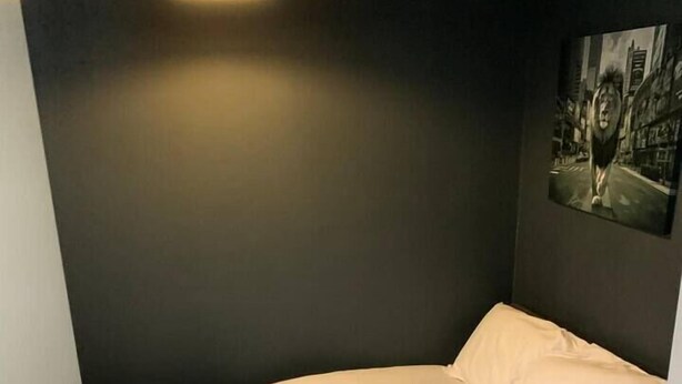 Quarto pequeno com uma cama de solteiro, parede escura, um quadro, abajur e chão de madeira.