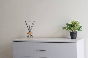 Ein weißer Schreibtisch mit einer Schublade, eine kleine Vase mit Schilfhalmen und ein Blumentopf mit weißen Blüten.