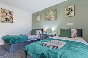 A room with two beds, each with a green bedspread, a nightstand with a lamp, and a wall with three framed pictures. - #134 Cozy Retreat Close to FarGo Village -Sleeps 7 (West Midlands)
