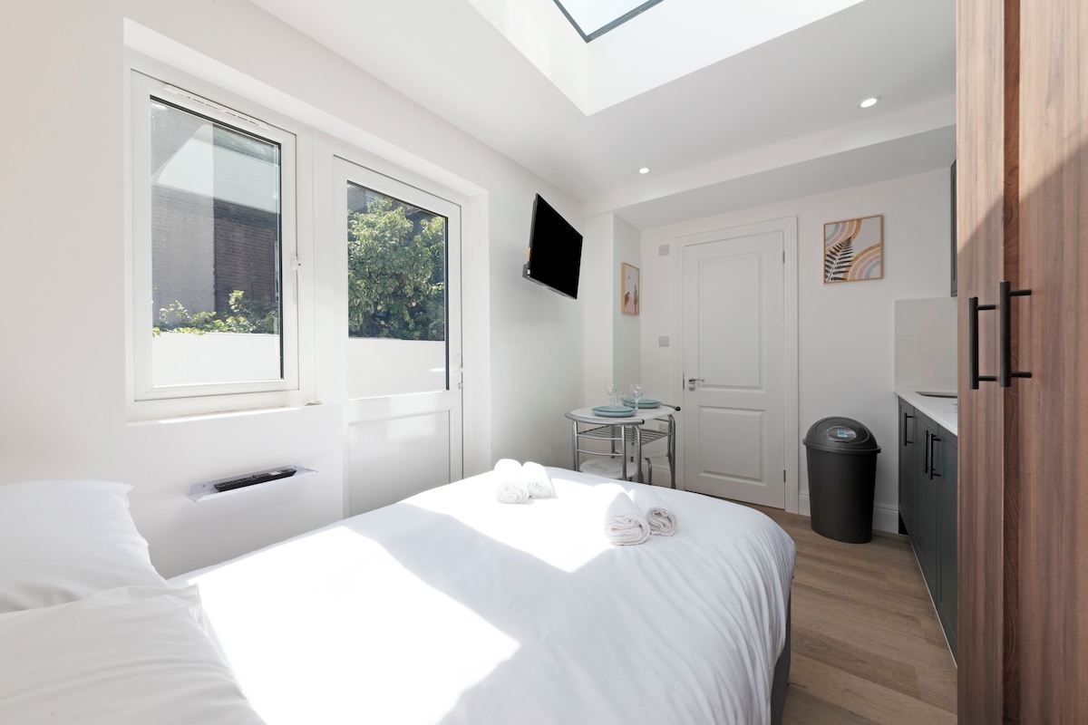 A modern bedroom with a skylight, a bed with white linens, a small round table with a chair, and a TV mounted on the wall.