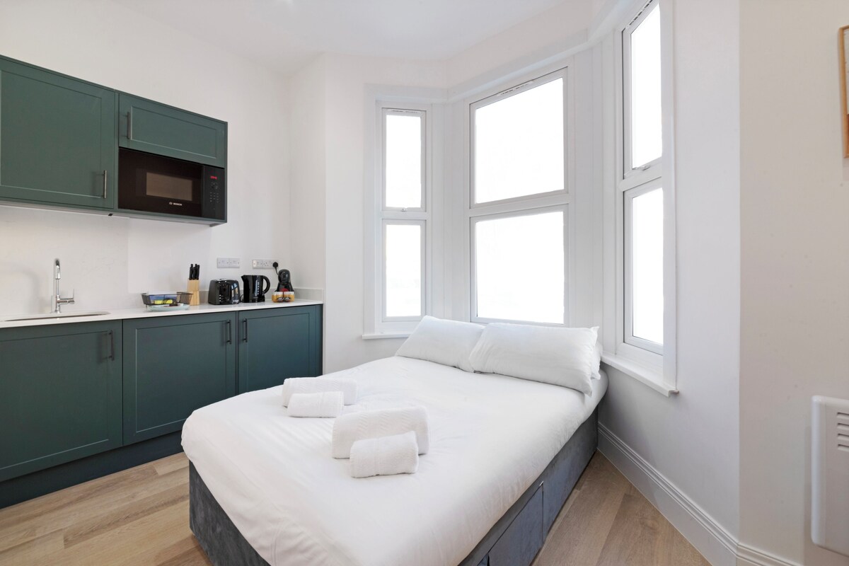 A modern bedroom with a large bed, a kitchenette with a microwave, and a window with multiple panes.