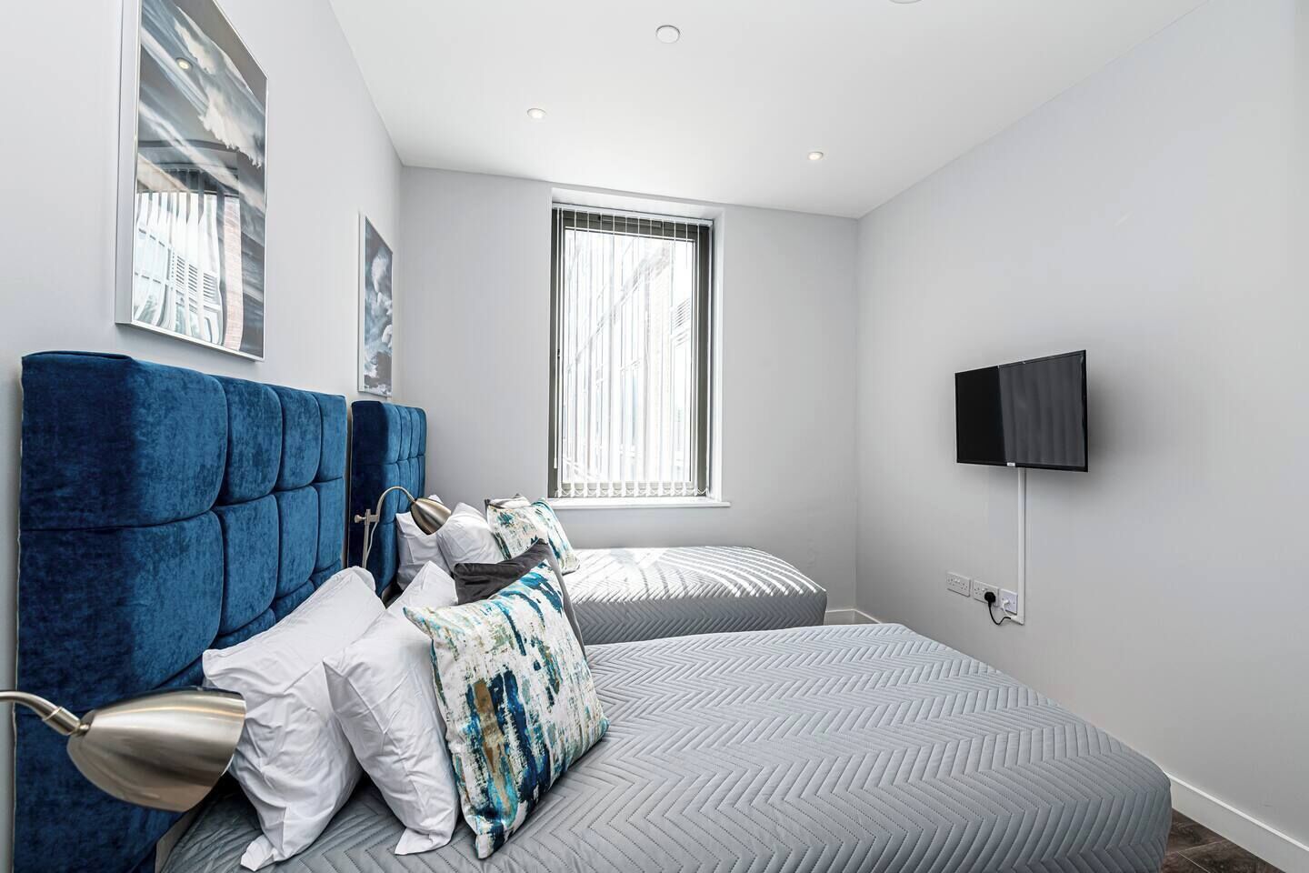 A modern bedroom with a large bed, a smaller bed, a TV, and a window with blinds.