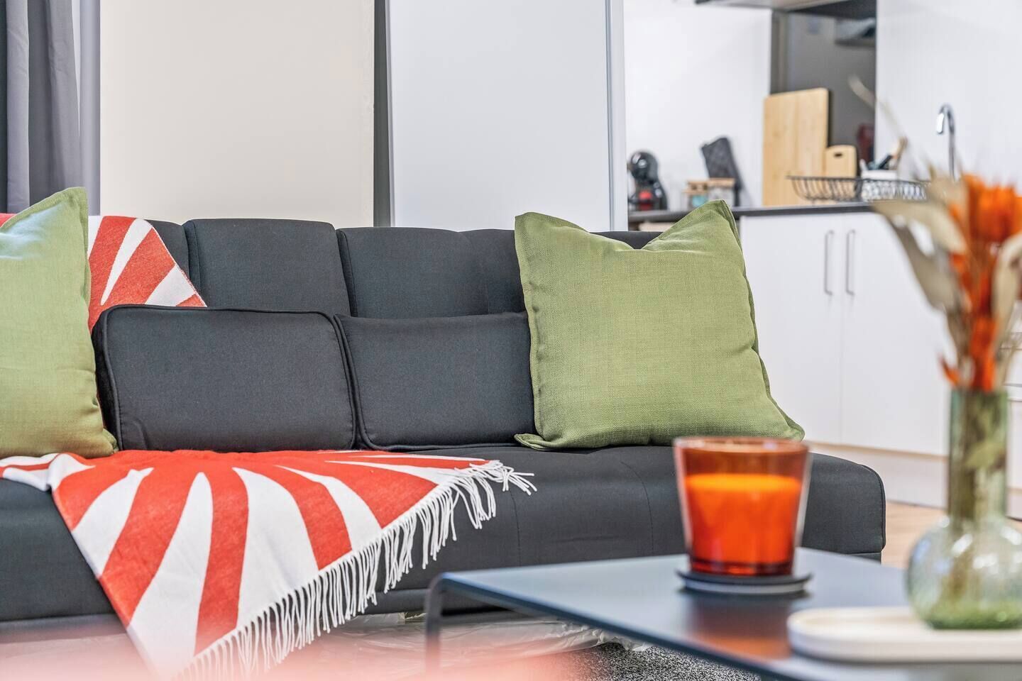 A modern living room with a black sofa, green and red cushions, a glass table, and a candle.