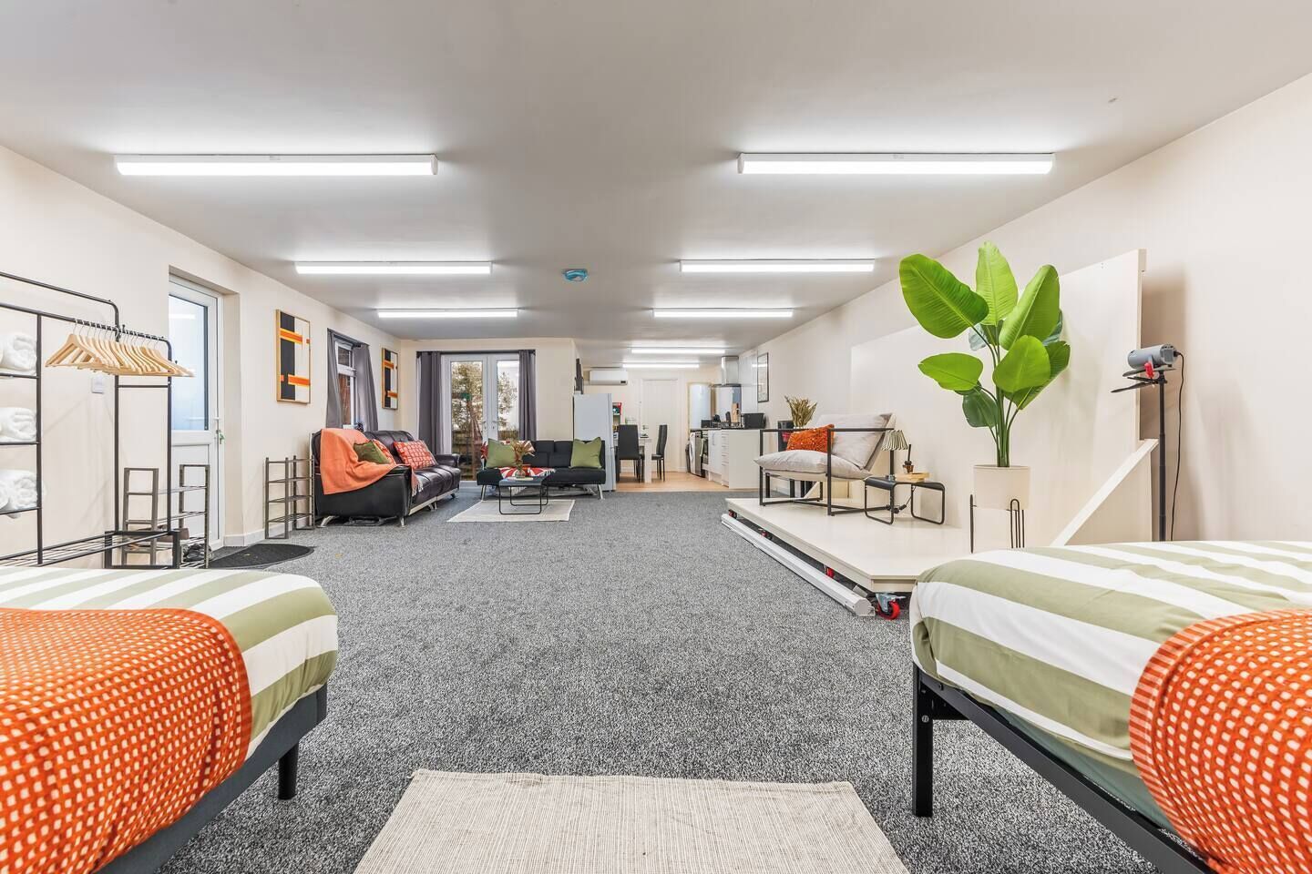 A modern, well-lit room with a sofa, two beds, a desk, and a plant.
