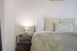 Ein ordentlich bezogenes Bett mit weißer Decke und zwei Kissen, ein Nachttisch mit Lampe und Fernbedienung.