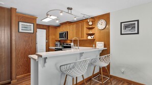 Una cucina con mobili in legno, un piano di lavoro bianco, un frigorifero in acciaio inossidabile, un forno a microonde, un lavandino e due sgabelli da bar.