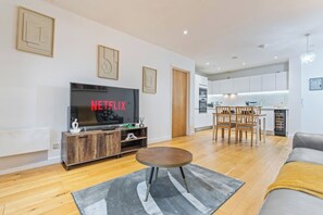 A modern living room with a flat-screen TV displaying 'NETFLIX,' a wooden entertainment unit, a round coffee table, and a dining area with wooden chairs and a kitchen in the background. - #178 Luxury Flat in the Heart of Birmingham-for 4 (Birmingham)