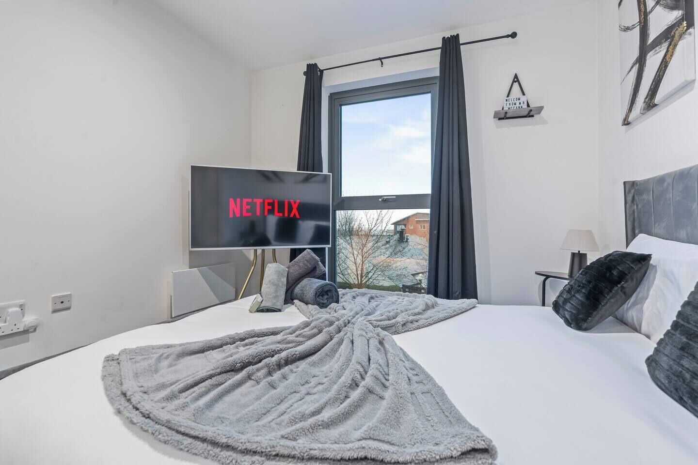 Une chambre d’hôtel avec un lit, un téléviseur affichant le logo « NETFLIX », une fenêtre avec des rideaux et une vue sur les bâtiments environnants.