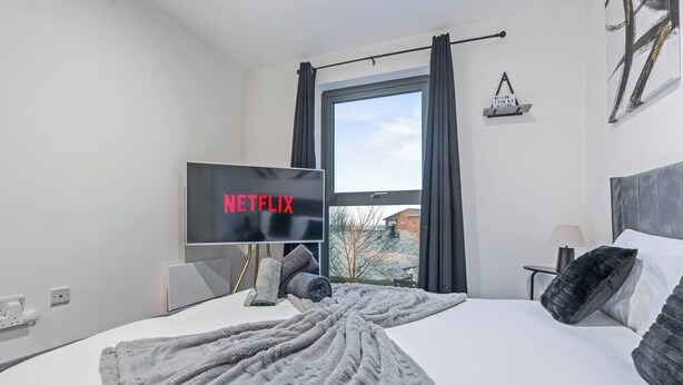 Quarto de hotel com uma cama, uma TV exibindo 'NETFLIX', uma janela com cortinas e vista para edifícios.