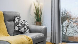 Uma sala de estar moderna com um sofá cinzento, um cobertor amarelo e uma almofada geométrica. Um vaso com planta está no chão ao lado do sofá.