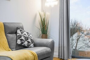 Ein modernes Wohnzimmer mit einem grauen Sofa, einem gelben Überwurf und einem geometrischen Kissen. Eine Topfpflanze steht auf dem Boden neben dem Sofa.