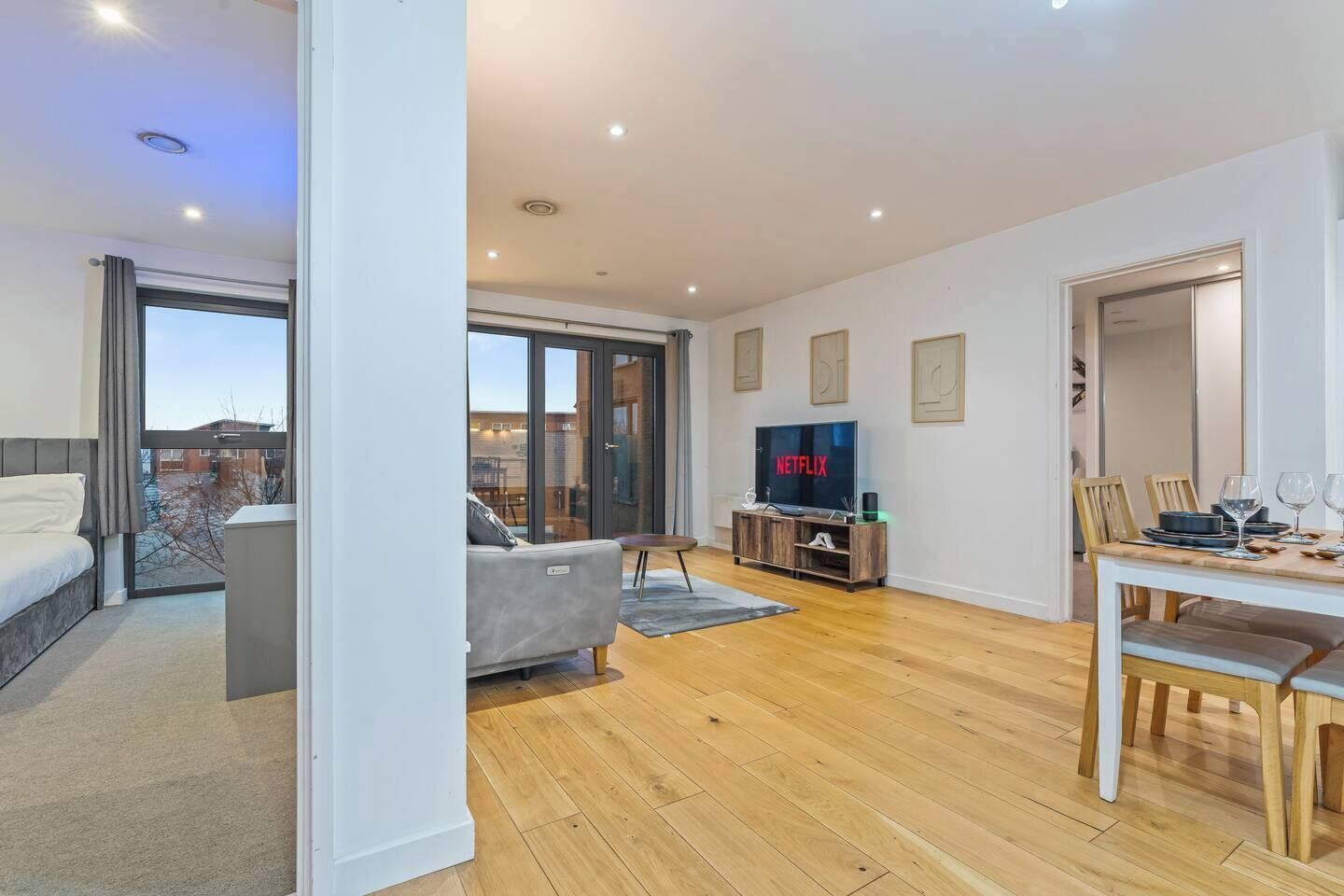Una sala de estar moderna con un sofá, una mesa de comedor preparada para dos personas y un televisor mostrando 'NETFLIX'.