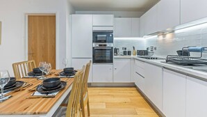 Uma cozinha moderna com armários brancos, uma mesa de jantar de madeira para quatro pessoas e um forno embutido.