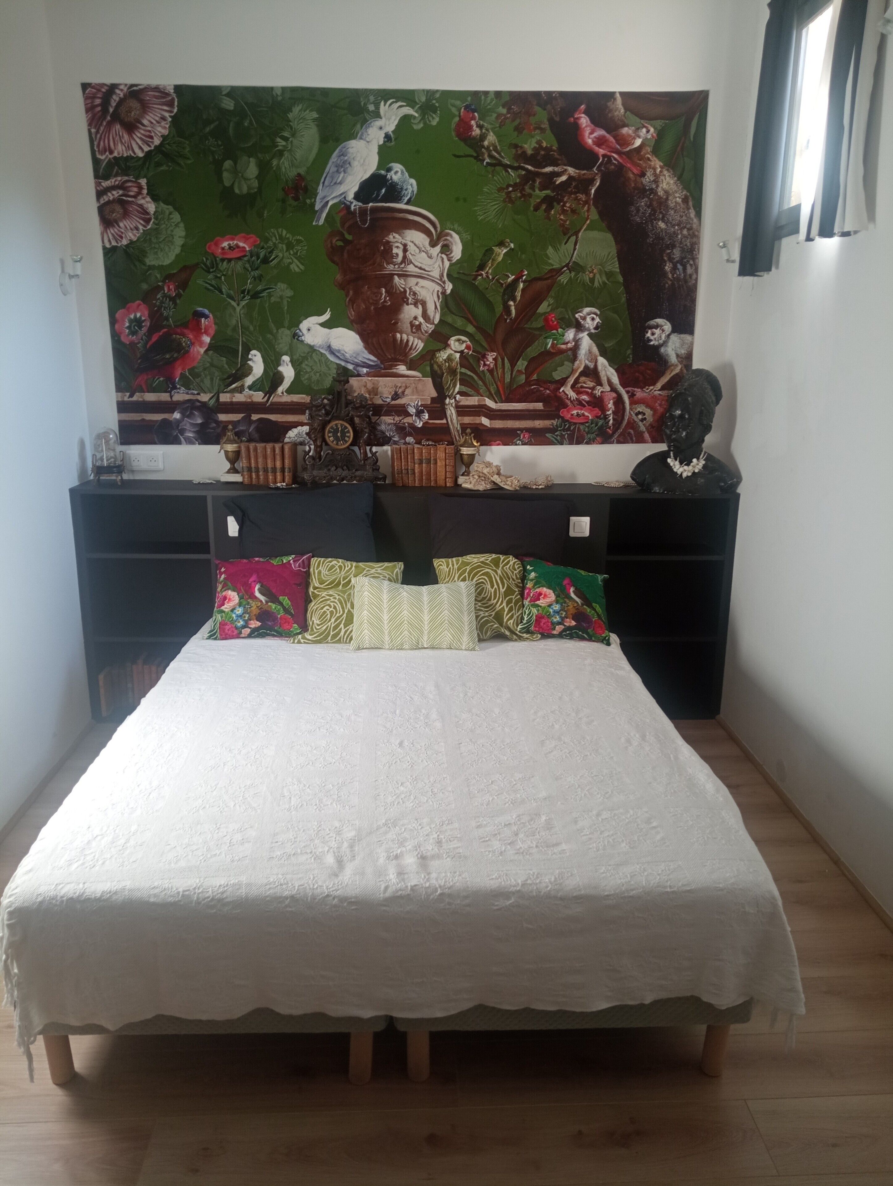 A bed with a white bedspread, a nightstand with a clock, and a wall tapestry with birds and flowers.