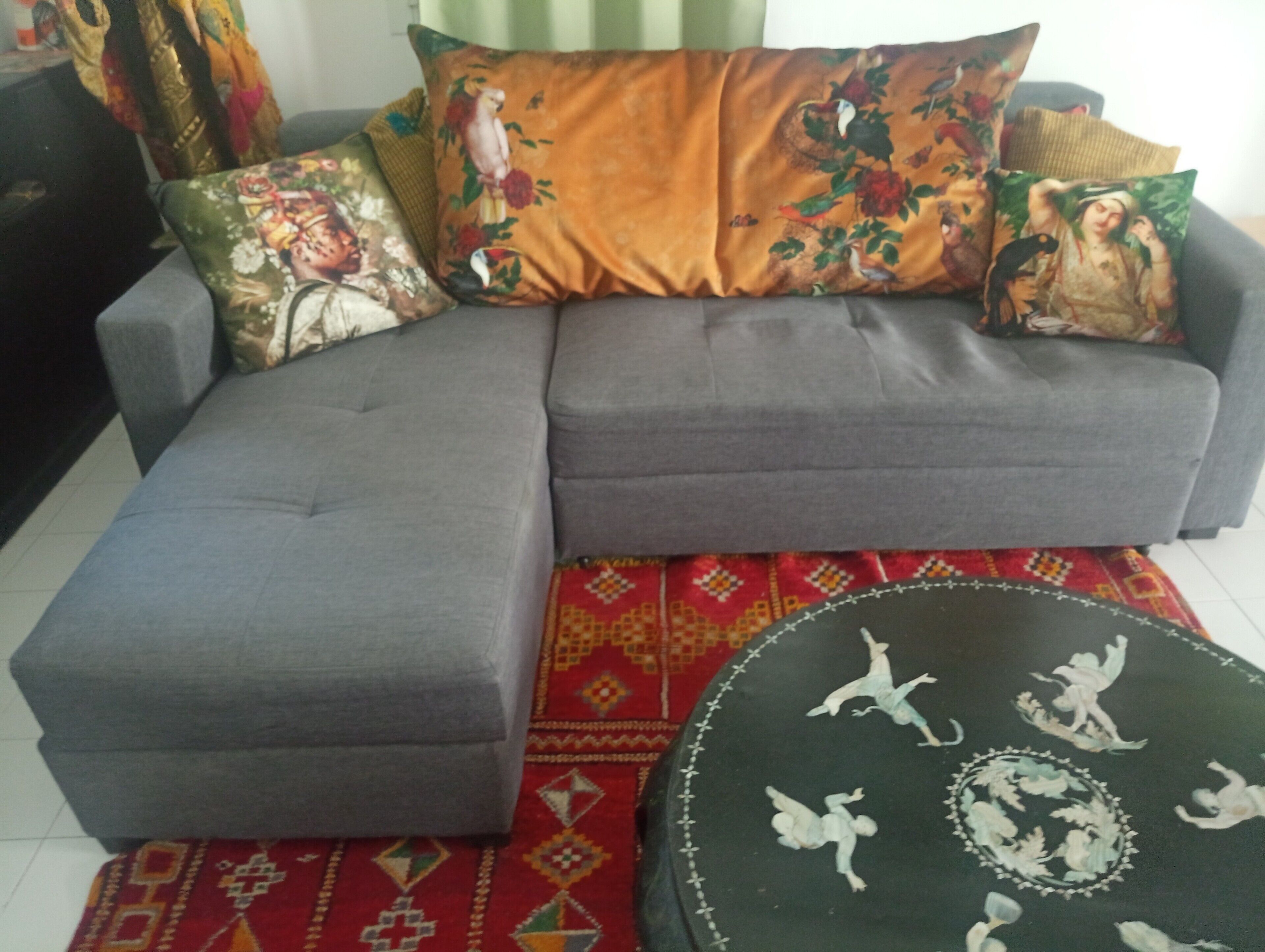 A grey sectional sofa with floral and nature-themed cushions, a patterned red rug, and a round ottoman with decorative figures.