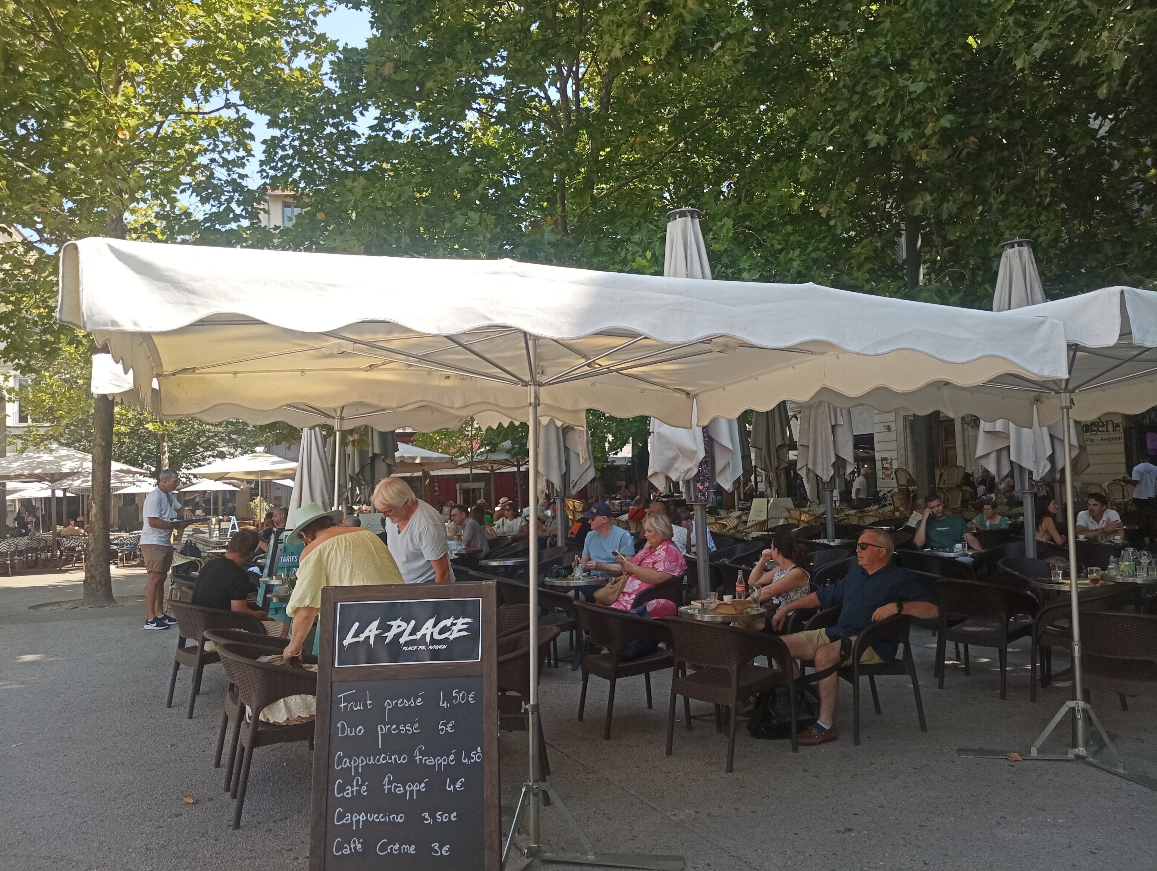 An outdoor café with tables and chairs under a canopy, a chalkboard menu, and people dining.