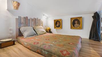 A bedroom with a wooden floor, a bed with patterned bedding, a nightstand with a lamp, and two framed portraits on the wall.
