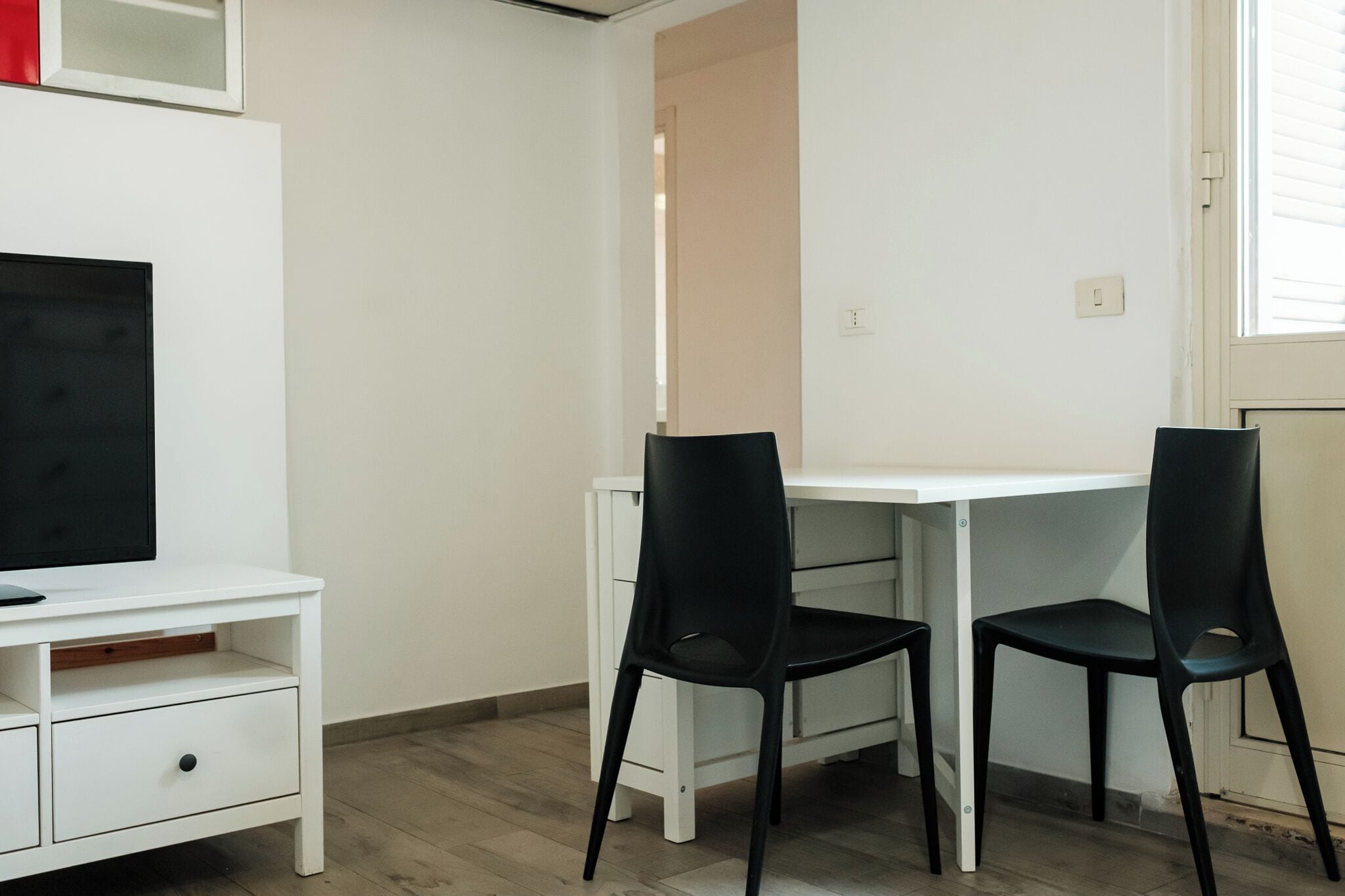 A small, clean room with a white desk, two black chairs, a television on a white stand, and a red object on the wall.