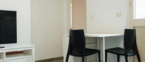 A small, clean room with a white desk, two black chairs, a television on a white stand, and a red object on the wall.