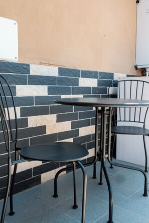 A patio area with a table and two chairs, a tiled wall with a stone pattern, and a whiteboard on the wall. - Terrace & Relax | Rome Collection (Roma)