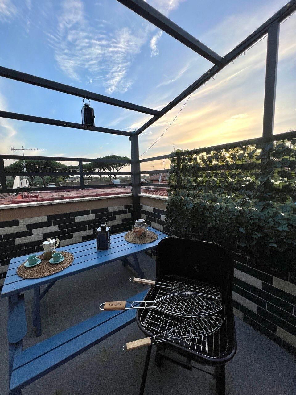 A rooftop patio with a barbecue grill, a table with a teapot and cups, and a view of the cityscape.