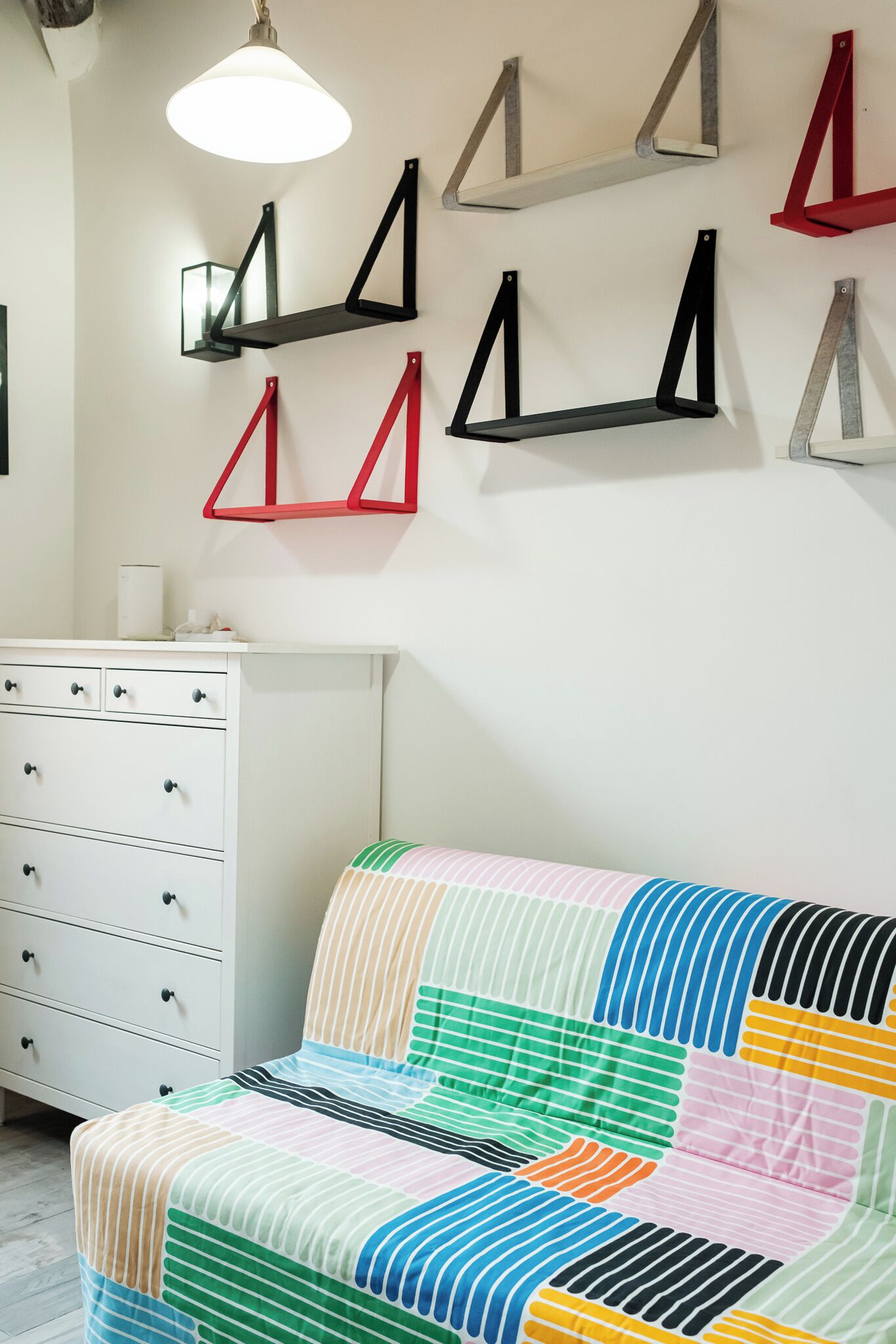 A room with a white dresser, a colorful striped sofa, and geometric wall shelves.