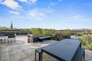 An outdoor patio with a table and chairs, surrounded by greenery and a view of a distant tower. - #179 Chic Birmingham City Escape - Sleeps 4 (West Midlands)