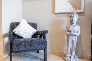 Una silla con tela texturizada y un cojín blanco, una estatua de una figura sentada, y una obra de arte enmarcada en la pared.