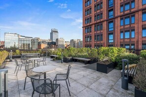 Eine Dachterrasse mit Sitzgelegenheiten im Freien, Blumentöpfen und Blick auf die Stadt.
